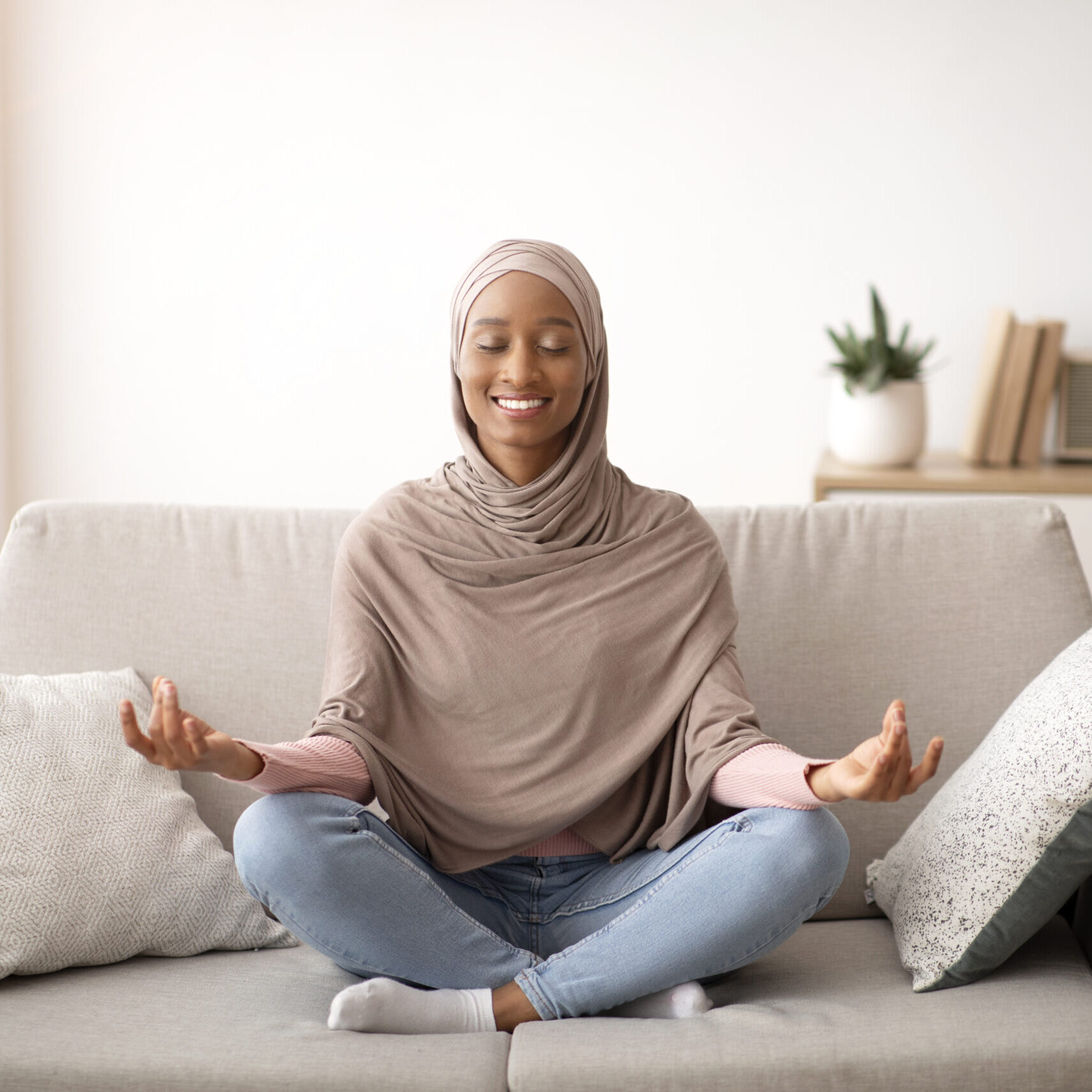 Stress management concept. Peaceful young lady in hijab meditating with closed eyes on sofa at home. Pretty Muslim woman sitting on couch in yoga pose, finding inner balance